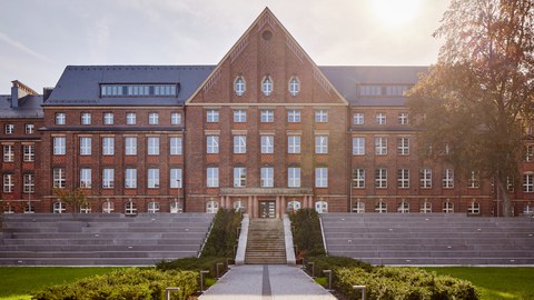 Foto vom Innenhof des Fritz-Förster-Baus. Es wurde frontal fotografiert und zeigt den begrünten Weg zum Hauptgebäude mit den großen Treppenstufen. Die Sonne steht hoch über dem Gebäude.