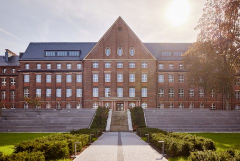 Foto vom Innenhof des Fritz-Förster-Baus. Es wurde frontal fotografiert und zeigt den begrünten Weg zum Hauptgebäude mit den großen Treppenstufen. Die Sonne steht hoch über dem Gebäude.