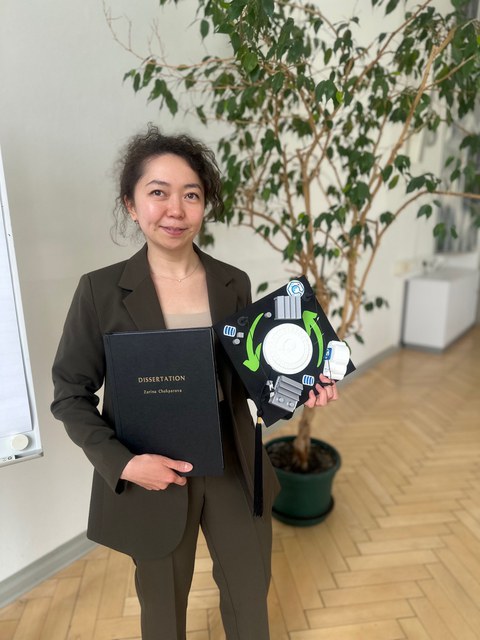 Zarina Chokparova hält in der rechten hand ihre Dissertation und in der linken Hand ihren persönlichen Doktorhut