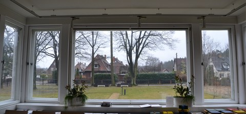 Foto zeigt den Blick aus dem Wintergarten des Wachmannshauses. Zu sehen ist eine Wiesenfläche mit angrenzenden Großgehölzen.