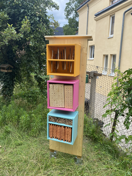 Bild des fertigen Insektenhotels in blau, pink und gelb gestrichen und auf einer Wiese aufgestellt