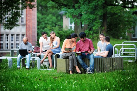 Das Foto zeigt mehrere Studierende draußen im Grünen. Sie sitzen auf einer geschwungenen Betonformation aus Tischen und Bänken und lernen.