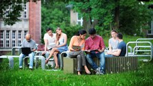 Das Foto zeigt mehrere Studierende draußen im Grünen. Sie sitzen auf einer geschwungenen Betonformation aus Tischen und Bänken und lernen.