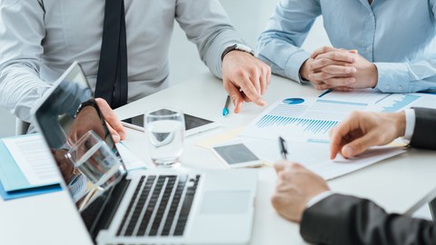 Auf dem Foto sieht man eine Gruppe, die gemeinsam in einem Meeting an einem Tisch sitzen. Auf dem Tisch steht ein Laptop und es liegen verschiedene Papiere vor den Personen. 