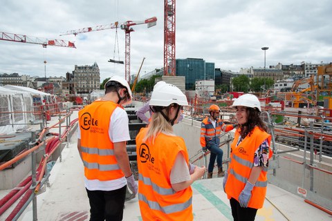 Mit Bauhelmen auf dem Kopf, stehen die Studierenden auf einer Baustelle