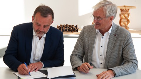 Die beiden Direktoren sitzen gemeinsam am Tisch und unterzeichnen einen Vertrag