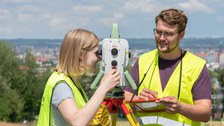 Zwei Studenten haben gelbe Leuchtwesten an, stehen auf einem Feld und schauen durch ein Messinstrument.