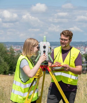 Zwei Studenten haben gelbe Leuchtwesten an, stehen auf einem Feld und schauen durch ein Messinstrument.