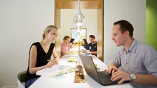 Foto von Studierenden in einer Cafeteria. Die Studenten sitzen mit Kaffee, Laptop und Buch an einem hohen Tisch und unterhalten sich.