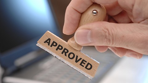 Das Foto zeigt die Hand einer Person. In der Hand hält diese einen Stempel mit dem Aufdruck "Approved".