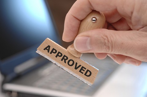 Das Foto zeigt die Hand einer Person. In der Hand hält diese einen Stempel mit dem Aufdruck "Approved".