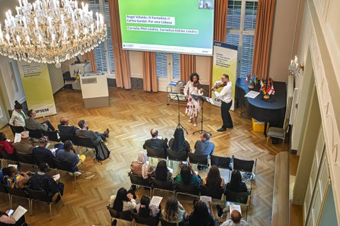 Cornelia Pfeil and Cornelius Köhler play violin in front of a backdrop featuring flags of various countries, with some audience members visible in the foreground.