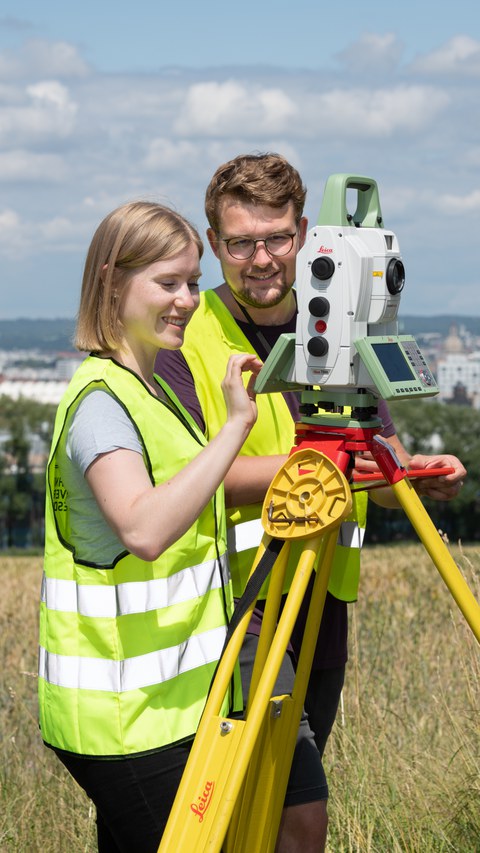 Geodätin und Geodät auf Feld, schauen durch Vermessungsinstrument Leica, machen Notizen