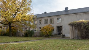 Drudebau front mit Baum im Herbstlaub, Wiese und Weg