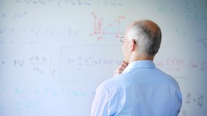 Ein älterer Mann steht nachdenklich vor einem mit Formeln beschriebenen Whiteboard. 