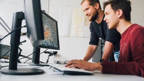 Two men are sitting at a desk and looking at grafics on a monitor