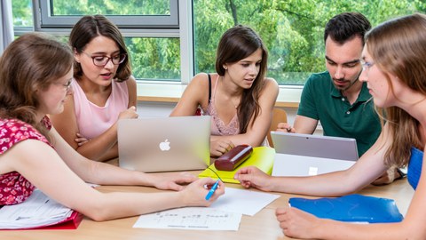 Four people are sitting at a table working together with paper, pen, and laptop.
