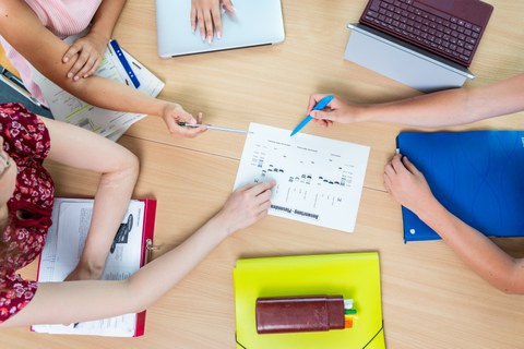 Students in a workshop session, students sitting at a table, only their hands are visible, in the middle is a sheet of paper they are discussing.