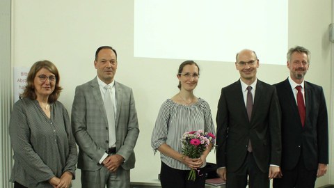 5 people are in a row. The woman in the middle holds a small bouquet of flowers in her hands.