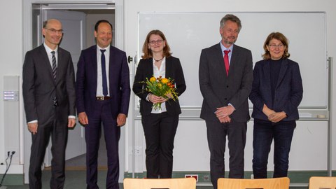 5 Personen in förmlicher Kleidung. Die Frau in der Mitte hält einen Blumenstrauß in der Hand.