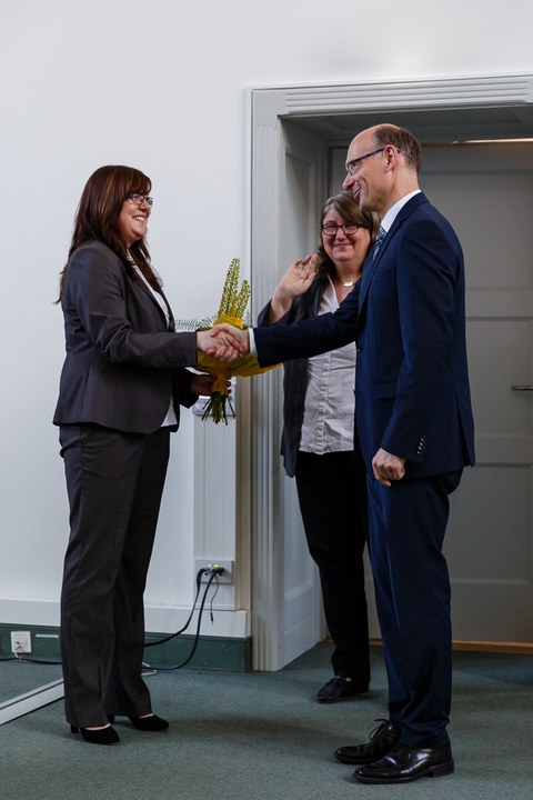 A man shakes hands with a woman. She holds a bouquet of flowers in her hand. In the background there is another woman.