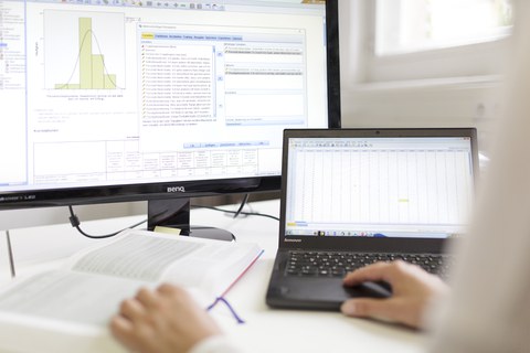 Zu sehen ist ein Bildschirm, auf dem SPSS zu sehen ist. Davor sitzt eine Person mit einem Laptop und einem Buch.  