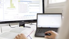 Zu sehen ist ein Bildschirm, auf dem SPSS zu sehen ist. Davor sitzt eine Person mit einem Laptop und einem Buch.  