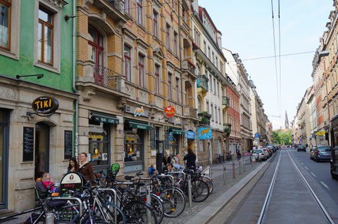 Es ist eine Straße in der Dresdner Neustadt zu sehen. Man sieht Straßenbahnschienen, Läden und viele Fahrräder  