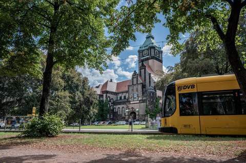 Foto vom Georg-Schumann-Bau und der Haltestelle davor mit einer Straßenbahn und den Fußgängern.