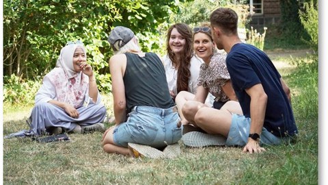 Es sind lachende Studierende zu sehen, die auf einer Wiese sitzen