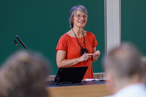 Zu sehen ist eine Frau mit einem roten Oberteil, die in einem Hörsaal einen Vortrag hält. Auf dem Pult sind Mikrofone und ein Laptop zu sehen. Im Hintergrund sieht man eine Tafel  