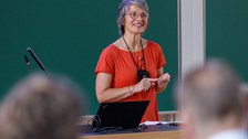 Zu sehen ist eine Frau mit einem roten Oberteil, die in einem Hörsaal einen Vortrag hält. Auf dem Pult sind Mikrofone und ein Laptop zu sehen. Im Hintergrund sieht man eine Tafel  