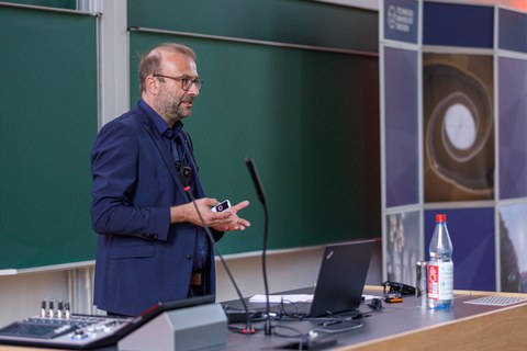 Es ist ein Mann in einem dunkelblauen Jacket zu sehen, der einen Vortrag hält. Man sieht ein Rednerpult und im Hintergrund eine Tafel und eine Stellwand