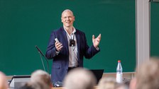 Es ist ein Mann mit weißem Hemd und blauen Jacket zu sehen, der in einem Hörsaal einen Vortrag hält. Im Hintergrund sieht man eine Tafel. 