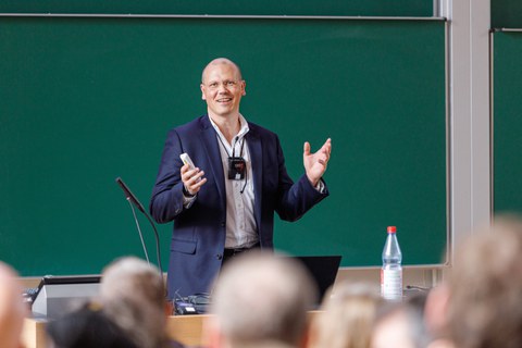 Es ist ein Mann mit weißem Hemd und blauen Jacket zu sehen, der in einem Hörsaal einen Vortrag hält. Im Hintergrund sieht man eine Tafel. 