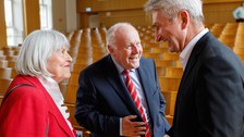 Zu sehen sind drei lachende ältere Menschen, die in einem leeren Hörsaal stehen