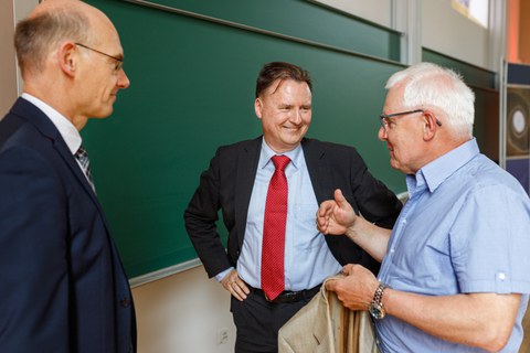 Zu sehen sind drei Männer, die vor einer Tafel stehen und sich freundlich unterhalten