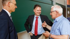 Zu sehen sind drei Männer, die vor einer Tafel stehen und sich freundlich unterhalten
