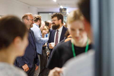Es ist ein gefüllter Gang im Hülße-Bau zu sehen, auf dem sich Menschen bei einer Kaffeepause miteinander unterhalten 