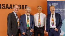 Es sind vier lachende Männer zu sehen, die alle die gleiche bunte Krawatte tragen. Im Hintergrund zu sehen ist eine orangefarbene Wand und ein blaues Rollup