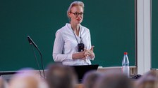 Zu sehen ist eine junge Frau, die ein Mikro umhängen hat und einen Vortrag in einem Hörsaal hält. Im Hintergrund ist eine Tafel zu sehen  