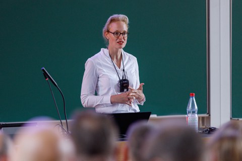 Zu sehen ist eine junge Frau, die ein Mikro umhängen hat und einen Vortrag in einem Hörsaal hält. Im Hintergrund ist eine Tafel zu sehen  