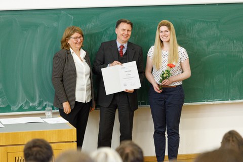 Es sind der Dekan, Prof. Strahringer und Dr. Katja Bley zu sehen. Es handelt sich um eine Preisverleihung. Katja Bley hält eine Blume in der Hand und bekommt eine Urkunde überreicht