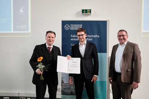 Es sind drei Männer zu sehen, die im Festsaal vor einem Rollup stehen. Es handelt sich um eine Preisverleihung. Der junge Mann in der Mitte bekommt eine Urkunde überreicht