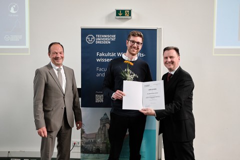 Es sind drei Männer zu sehen, die im Festsaal vor einem Rollup stehen. Es handelt sich um eine Preisverleihung. Der junge Mann in der Mitte bekommt eine Urkunde überreicht