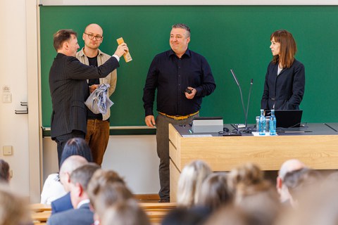 Zu sehen sind vier Menschen vor einer Tafel bei einer Preisverleihung  