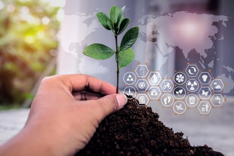 Im Vordergrund ist etwas Erde zu sehen und eine kleine grüne Pflanze. Im Hintergrund sieht man eine Weltkarte und verschiedene Icons, die für Umwelt und Energie stehen 