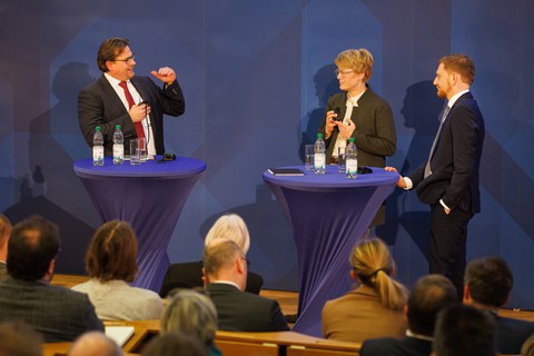 Die Podiumsdiskussion startet mit Veronika Grimm und Michael Kretschmer und wird von Prof. Leßmann moderiert.
