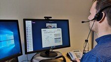 Es ist ein junger Mann zu sehen, der mit Headset auf dem Kopf vor einem Rechner sitzt und eine Videokonferenz verfolgt