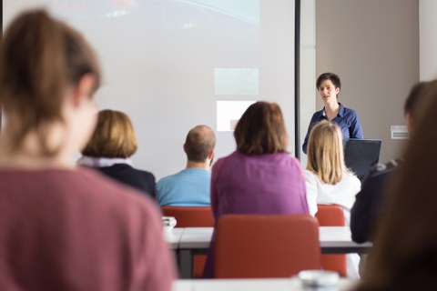 Das Foto zeigt eine Frau bei einer Präsentation vor vielen Personen